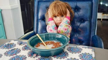 A little girl sitting in front of a half empty cereal bowl with her hands covering her ears