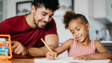 Dad smiling beside his daughter helping her with homework.