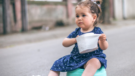 toddler girl sitting on a potty on the street holding toilet paper