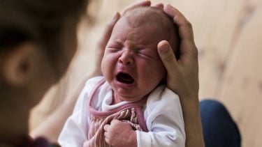 A newborn baby cries; their head is being cradled by an adult's hands