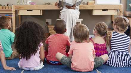 Little kids sitting in a classroom as a teacher reads a book