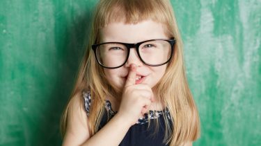 Child wearing glasses standing in front of a chalkboard with her finger placed in front of her mouth
