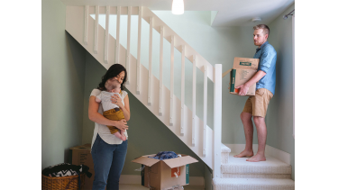 Husband carrying box upstairs while mother holds baby in her arms at new home