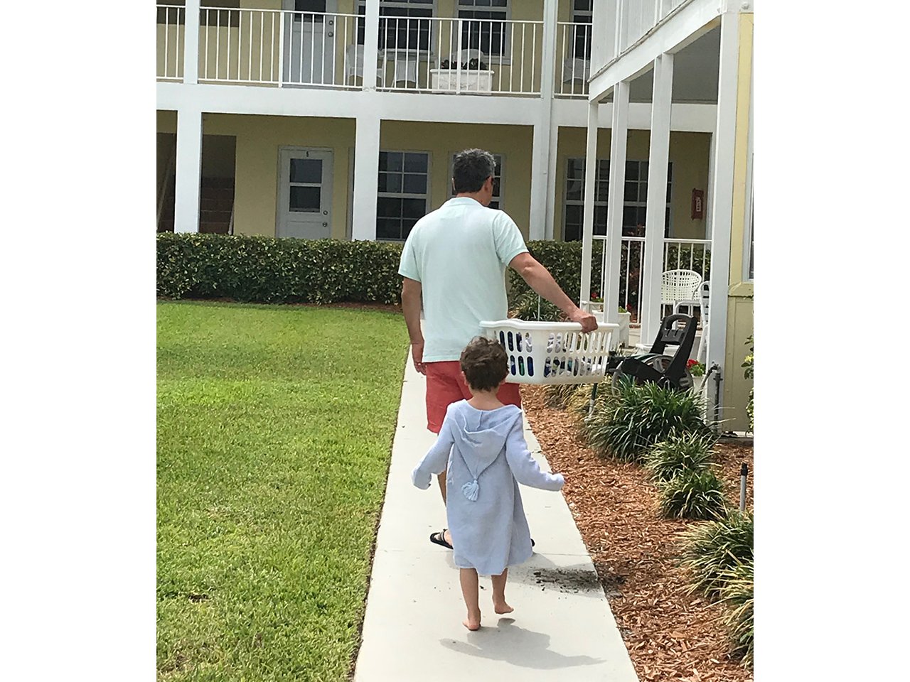 A dad holding a laundry basket with a little boy walking behind him up a pathway at a beach resort