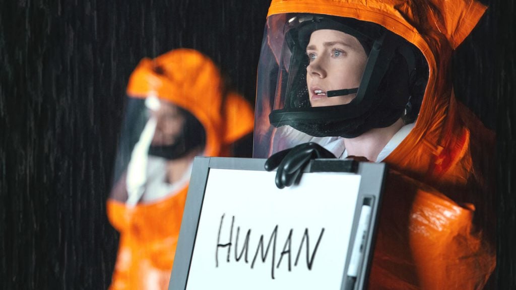 Promo image for Arrival showing a woman in a hazmat suit with a whiteboard that says human