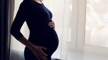Pregnant women in a blue body con dress holding her belly by a sheer-curtain window.