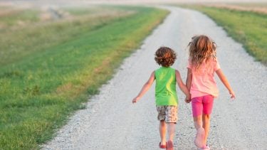 Older girl and younger boy holding hands as they walk along a gravel walkway.