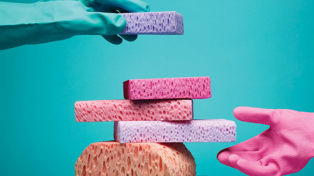 A blue dishwashing glove places a purple sponge on top of a stack of various pink sponges. A pink dishwashing glove is hovering near the bottom of the sponge tower.
