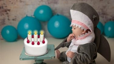 a baby dressed in a shark costume is sitting on the floor in front of a mystery cake.