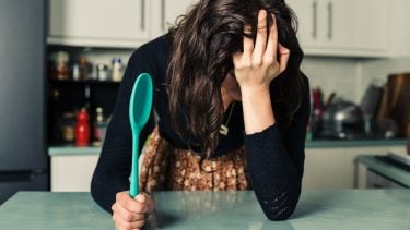 A sad young woman is standing in a kitchen with a spoon in her hand