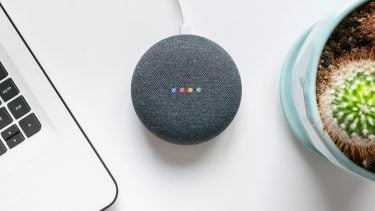 Google Home in listening mode; on a white desk beside a computer and cactus plant