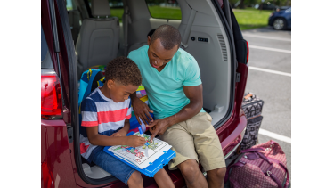 A father and son sitting in the back of an open trunk of a car, colouring with Crayola colour wonder markers, packed bags on the floor beside them