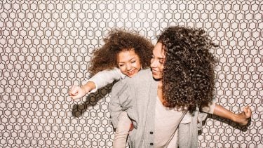 Stock image of a mother giving her daughter a playful piggy back in front of a decorative wall