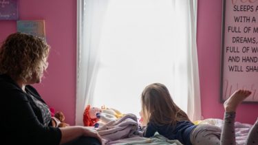 Adoptive mother watches adoptive daughter as she gazes out her bedroom window