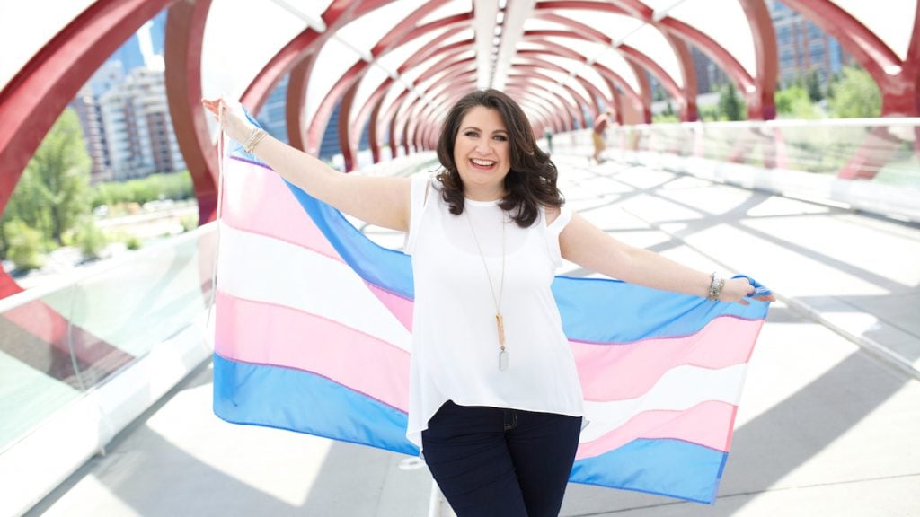 Tammy Plunkett smiles as she holds a transgender flag behind her.