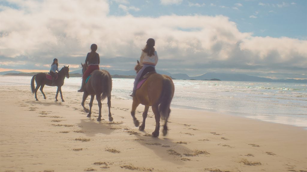 Promo image for Free Rein showing horseback riders riding on the beach