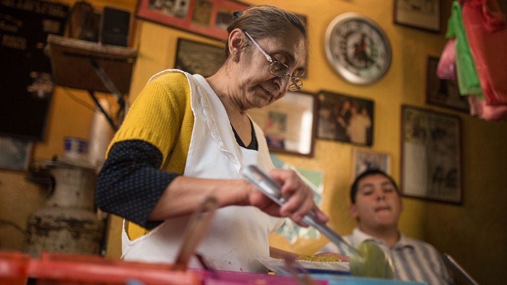 Promo image for Taco Chronicles showing an older woman cooking food