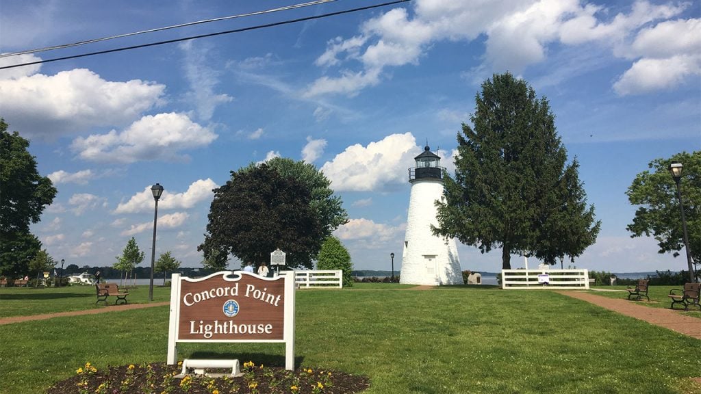 A lighthouse against a blue sky with clouds with a sign that says "Concord Point Lighthouse"