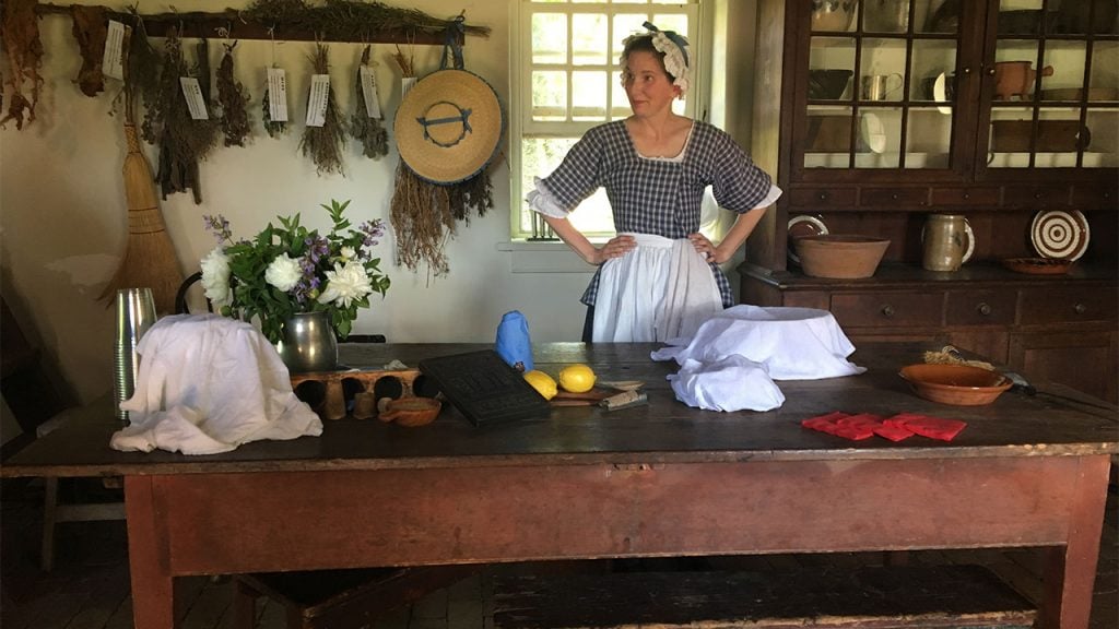 A woman in a period costume standing in a colonial era kitchen 