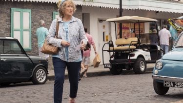 Meryl Streep in a t-shirt, button-up shirt and hat walking on a brick street