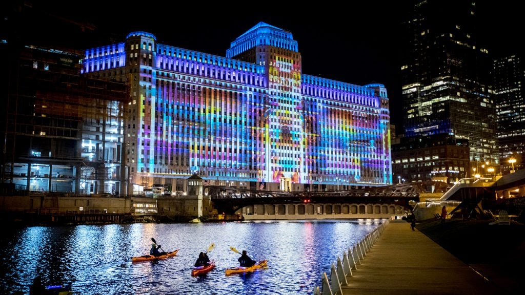 Kayakers paddle in front of theMART in chicago while art is projected onto the building's façade