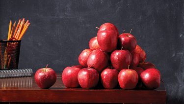 Teachers desk in front of a black board with a pile of red apples on it