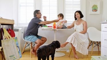 a man and woman sitting at a table feeding a toddler on the table while their dog stands by