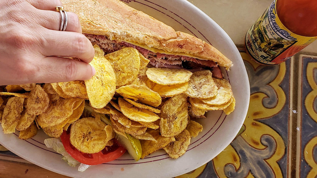 hand reaching in to eat plaintain chips of plate with Cuban sandwich