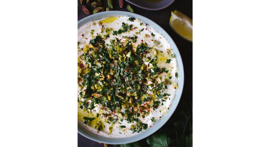 Bowl of labneh topped with oil, lemon, herbs and pistachios.
