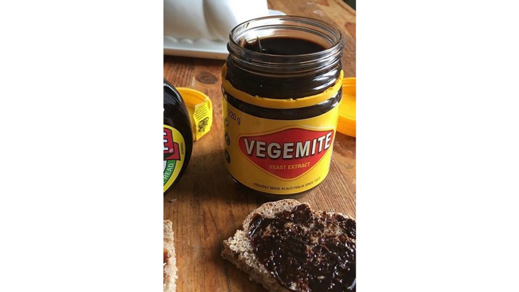 Open jar of Vegemite beside piece of bread