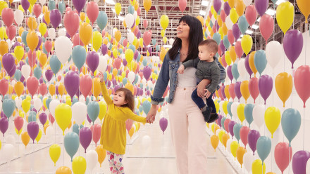 A mom, baby and toddler daughter having fun in a hallway of balloons and mirrors.
