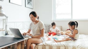 Young mother working at home while her two daughters playing on the bed