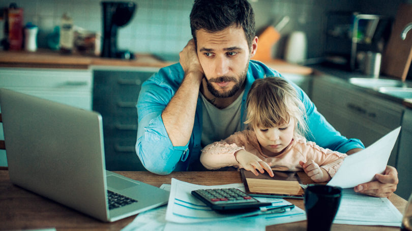 Stressed dad at the kitchen table looking at his computer and a bunch of papers with his daughter in his lap