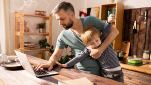 Dad carrying squirmy kid in the kitchen while working on a laptop