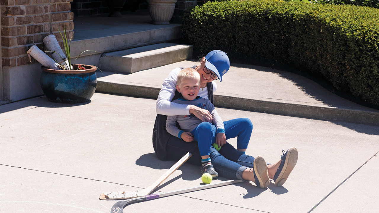Mom sitting on driveway with kid in lap and hockey sticks nearby