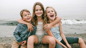 three kids sitting by the water together hugging and laughing