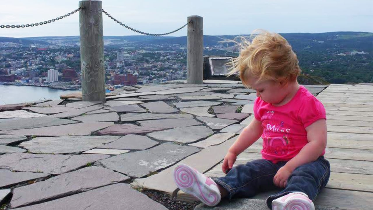 a toddler sitting on the ground with a view of downtown St. John's in the background