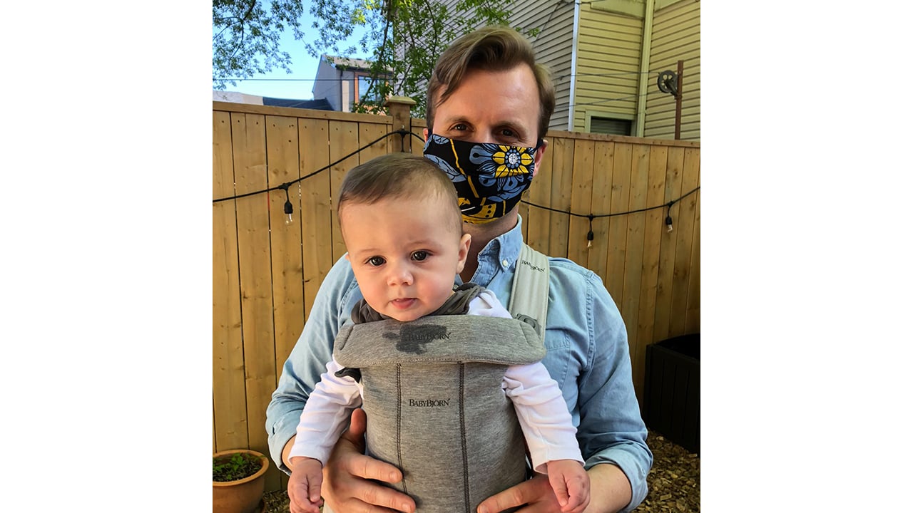Joe carrying his son Jude in a baby carrier while wearing a cloth face mask
