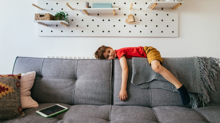 kid lying on back of couch looking bored with tablet laying nearby