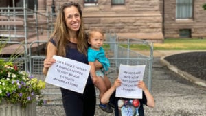 single mom with her babies holding protest signs that say I am a single parent and cannot afford to quit my job to teach my kids at home