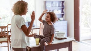 Happy little boy giving high-five to mother at home. Young woman is standing while son sitting on table. They are wearing casuals.
