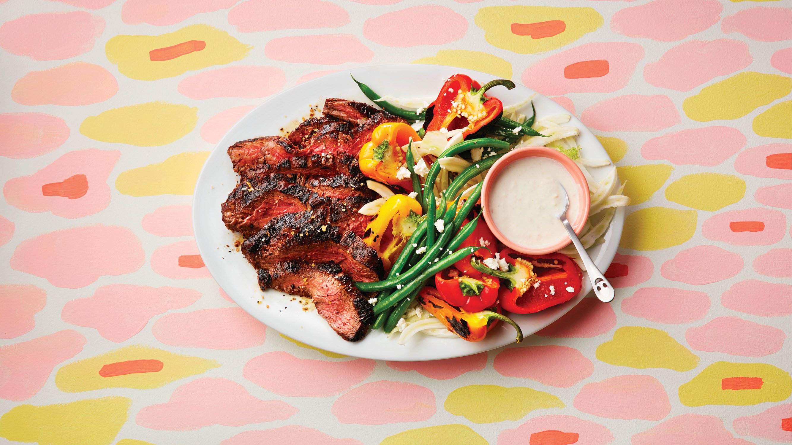 late summer recipe of plate with grilled flank steak, green beans, peppers and a yogurt dipping sauce on a patterned table cloth