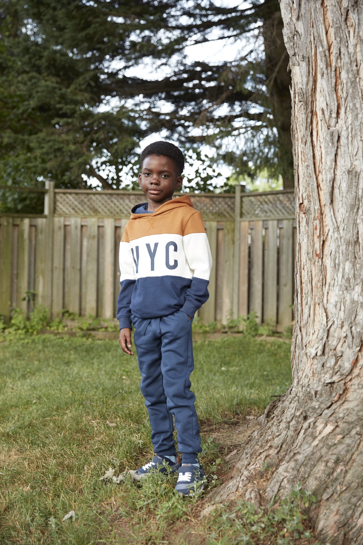 Eight-year-old wearing joggers and a hoodie in backyard beside tree