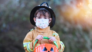 Photo of a little kid dressed for Halloween as a firefighter wearing a facemask and clutching a pumpkin bucket