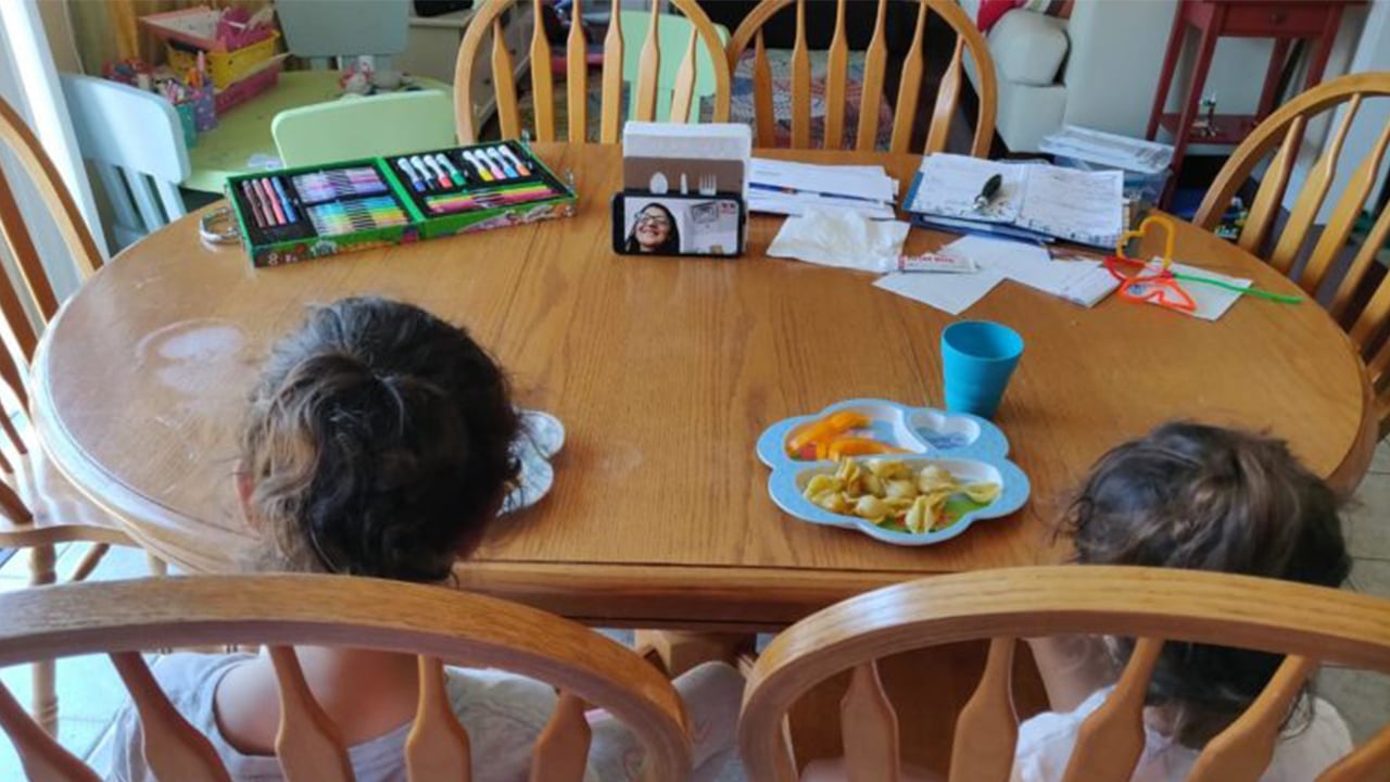 Photo of two kids sitting at the ktichen table eating while video chatting with a grown up on a phone