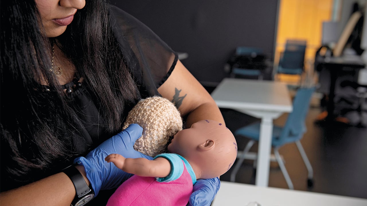 a woman training to be a midwife demonstrates breastfeeding with a baby doll and fake breast