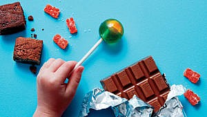 a kid's hand holding a green lollypop alongside brownies, gummies and a chocolate bar