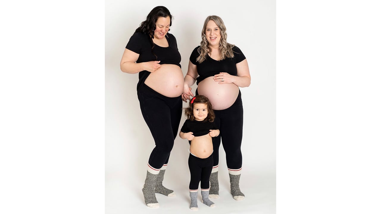 Photo of a lesbian couple with double pregnancies showing off their baby bumps while their first child shows off their own belly