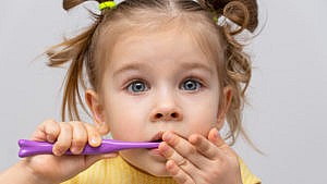 a nervous preschool-aged girl in a yellow shirt holds a purple toothbrush to her mouth