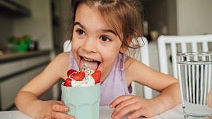 photo of a young kid joyously licking ice cream in a cup while sitting at the dinner table